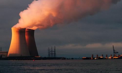 A power plant on a sea shore with smoke emerging from the towers. 